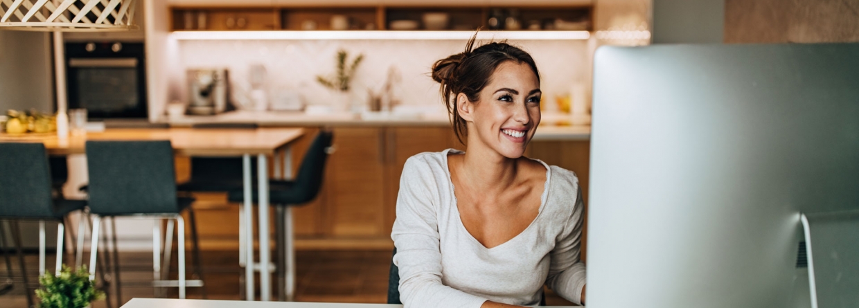 Een vrolijke vrouw die thuis aan het werk is achter de computer met op de achtergrond de keuken en eettafel